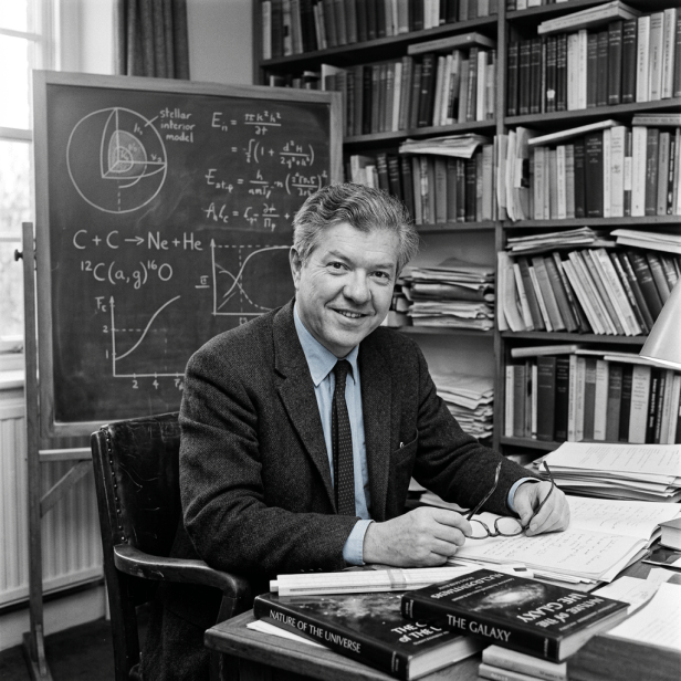 Scientist seated at desk with astrophysics books and chalkboard of equations